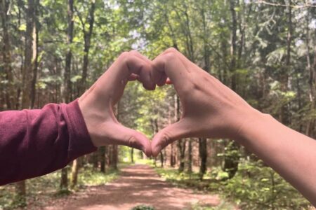 Zwei Hände formen gemeinsam ein Herz vor einem Waldweg – Zeit zu zweit als Paar
