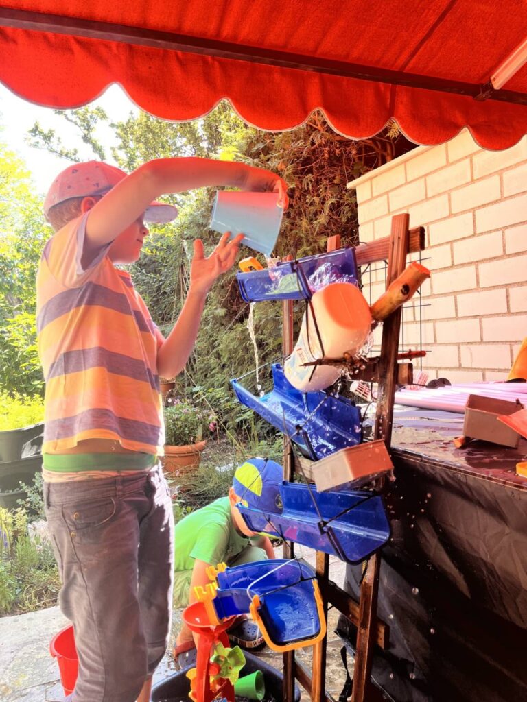 Kind gießt Wasser in selbstgebaute Wasserfall-Bahn – Wasserspiele bei Hitze im Garten
